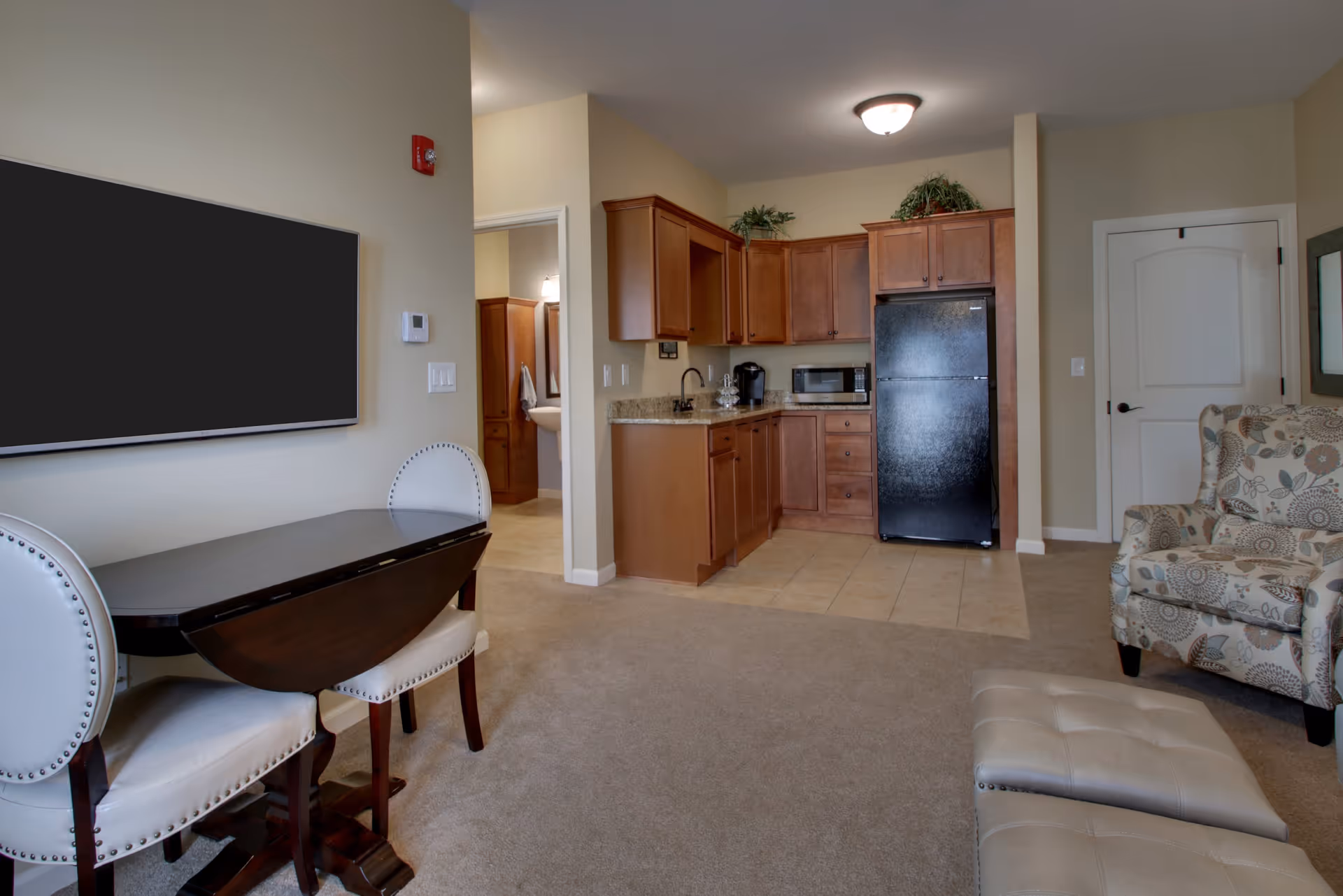 Interior view of a senior living facility room featuring a small kitchen area with wooden cabinets, a black refrigerator, microwave, and coffee maker. Adjacent to the kitchen is a dining area with a dark wooden table and two white upholstered chairs. A patterned armchair and a beige ottoman are visible in the living space. The room has beige walls and carpeted flooring with tile in the kitchen area. A doorway leads to a bathroom with a sink and mirror visible.