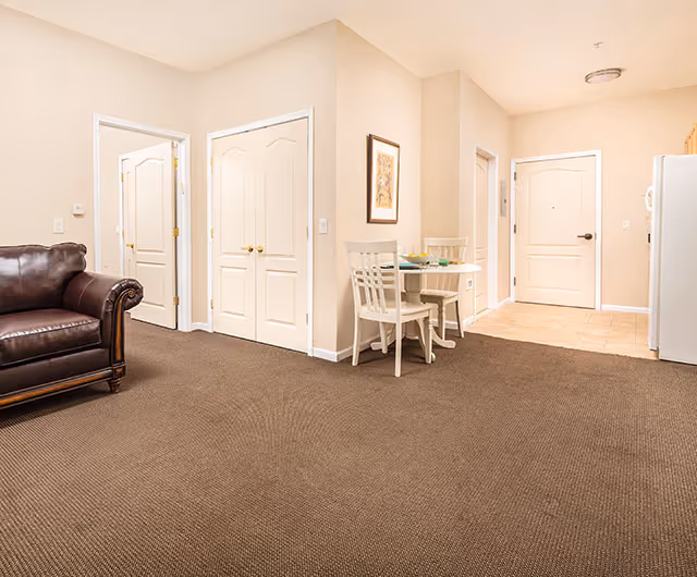 Interior view of a senior living facility room with beige walls and carpeted floor. There is a dark brown leather armchair on the left, a small round dining table with two white chairs in the center, and a white refrigerator on the right. Several white doors are visible, including a double-door closet and an entrance door with a tiled floor area near it. A framed picture hangs on the wall above the dining table.