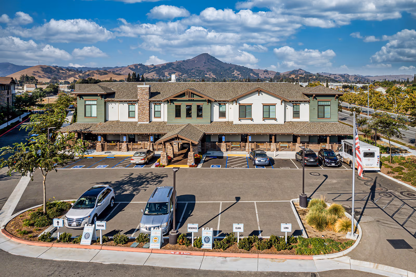 Front exterior of a two-story senior living building with a parking lot, an American flag, and hills in the background.