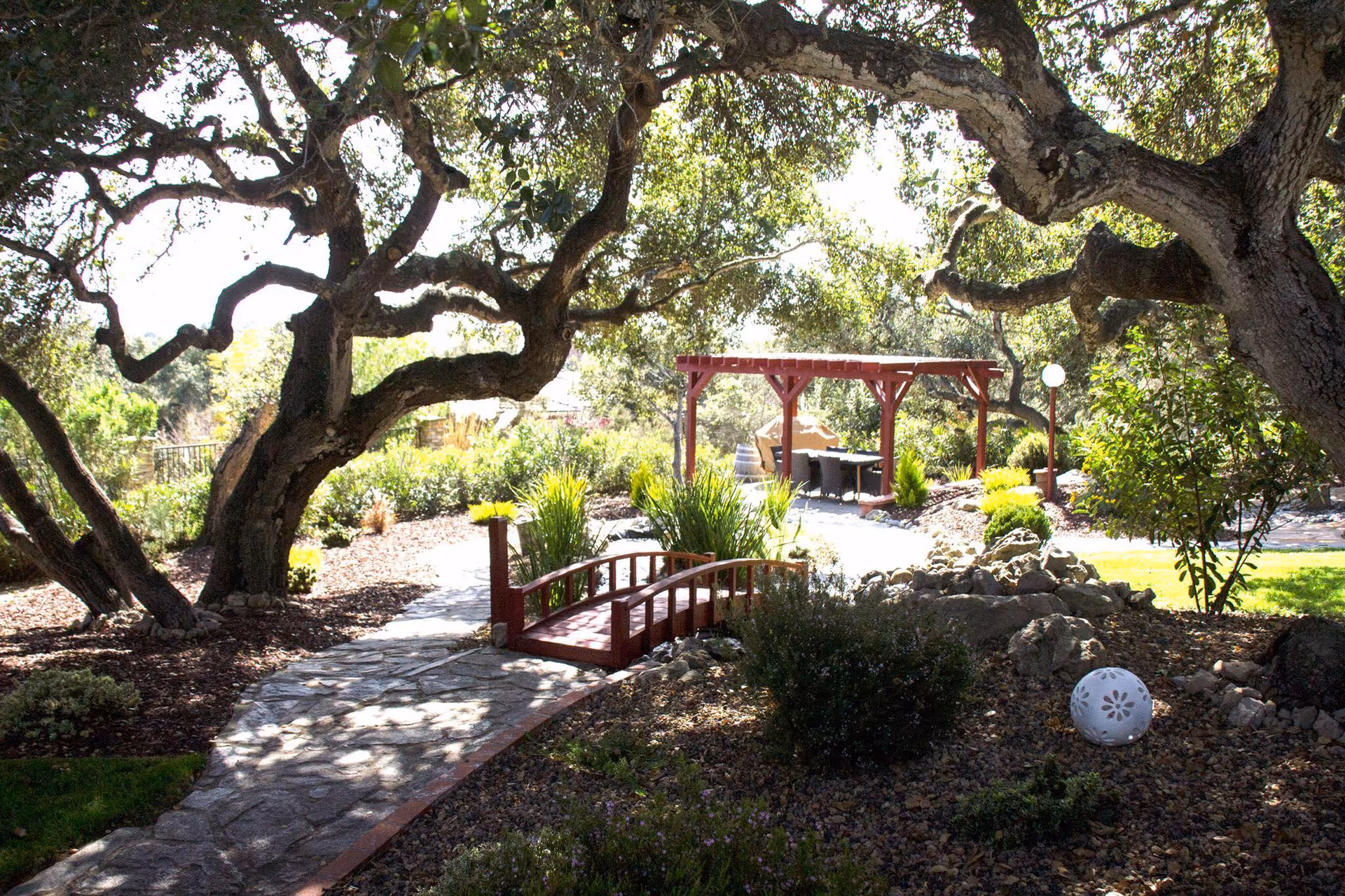A serene outdoor garden area with large, mature trees providing shade over a stone pathway. A small red wooden bridge crosses a dry rock bed, leading to a covered seating area with a table and chairs under a pergola. Various shrubs and plants surround the pathway and seating area, creating a peaceful natural environment.