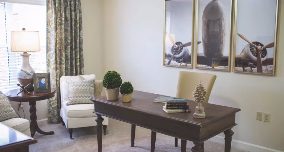 A cozy office or study room with a wooden desk holding books, two small potted plants, and a decorative tree. Behind the desk is a beige chair. To the left, there is a white armchair with patterned cushions next to a round wooden side table with a lamp and a framed photo. The wall features a three-panel artwork of an airplane front view. The room has light-colored walls and carpeted floor.
