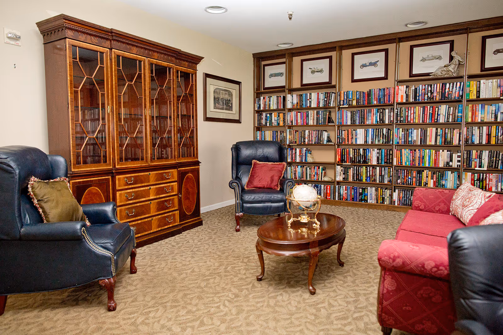A cozy library room with a large wooden bookshelf filled with books along the back wall. There are two dark blue leather armchairs with decorative pillows, a red upholstered sofa with patterned cushions, and a wooden coffee table with a decorative globe on it. A wooden cabinet with glass doors and drawers is against the left wall, and framed pictures hang on the walls. The room has beige carpet and neutral-colored walls.