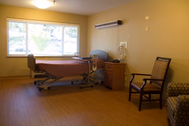 A simple room with a hospital-style bed covered with a brown blanket, positioned near a large window with blinds. Next to the bed is a small wooden nightstand with a telephone and medical equipment mounted on the wall above it. There is a wooden chair with armrests and a patterned cushion, as well as a patterned armchair partially visible on the right side. The floor has a wood-like finish and the walls are painted beige.