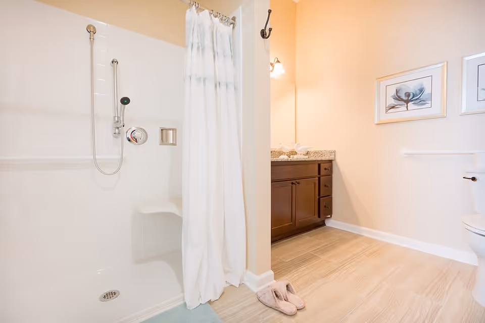 A clean and bright bathroom featuring a walk-in shower with a white curtain, a built-in shower seat, and a handheld showerhead. The bathroom also includes a wooden vanity with a granite countertop, a toilet, and two framed floral artworks on the wall. A pair of pink slippers is placed on the light wood floor near the shower.