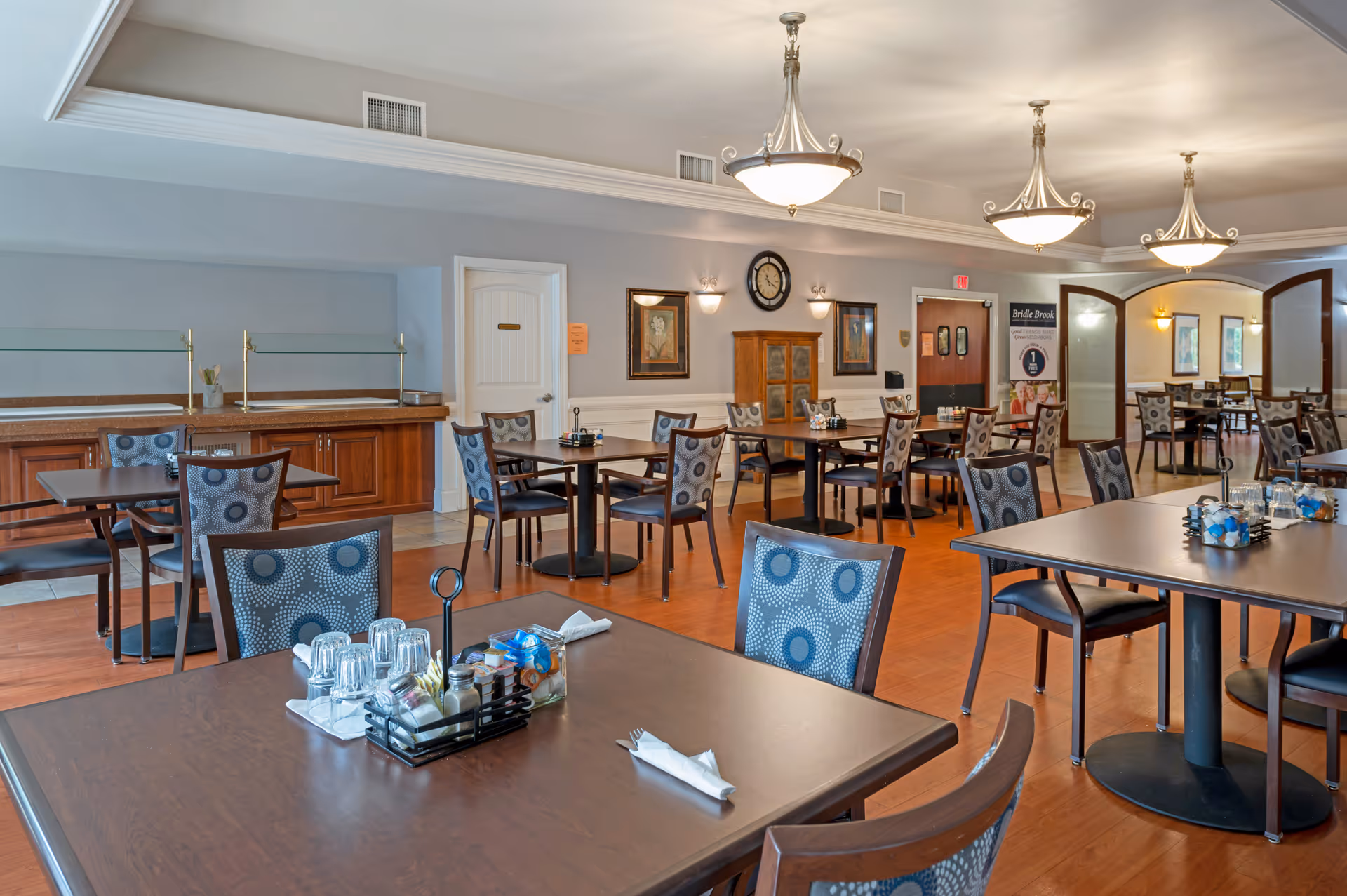 A spacious dining room in Bridle Brook Assisted Living & Memory Care Community featuring multiple dark wood tables and chairs with patterned upholstery. The room has warm wooden flooring, soft lighting from ceiling fixtures, and decorative wall art. There is a buffet counter along one wall and a clock mounted above a wooden cabinet.