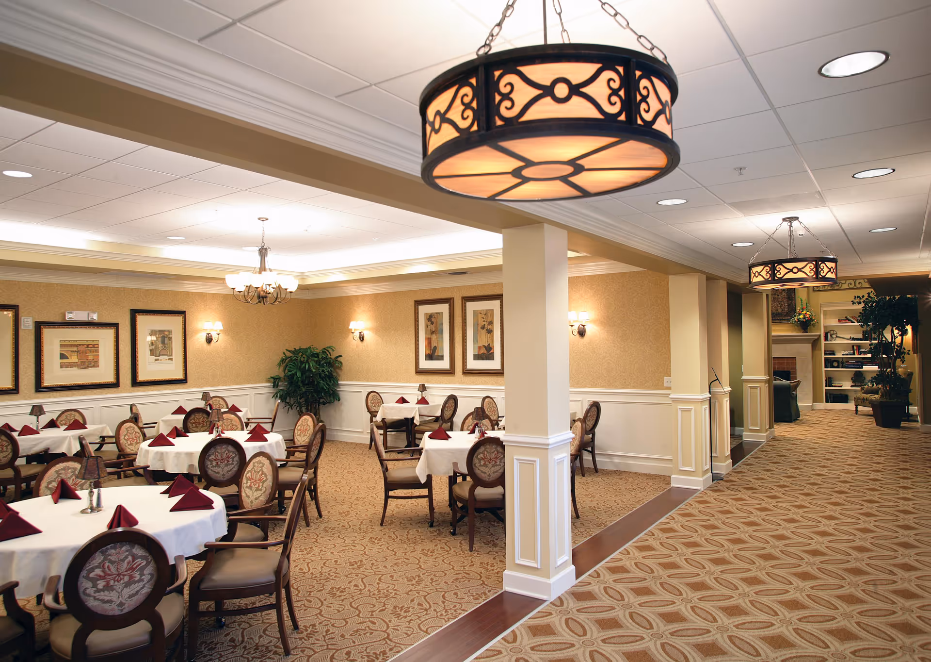 A well-lit dining room in a senior living facility with round tables covered in white tablecloths and set with folded burgundy napkins. The room features elegant upholstered chairs, framed artwork on the walls, decorative wall sconces, and a large potted plant in the corner. The adjacent hallway has patterned carpet, hanging light fixtures, and leads to a cozy seating area with bookshelves and plants.