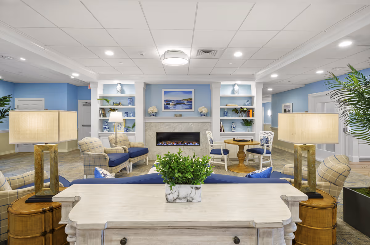 Well-lit communal living room with upholstered chairs and sofas facing a fireplace, built-in shelves, lamps and potted plants.