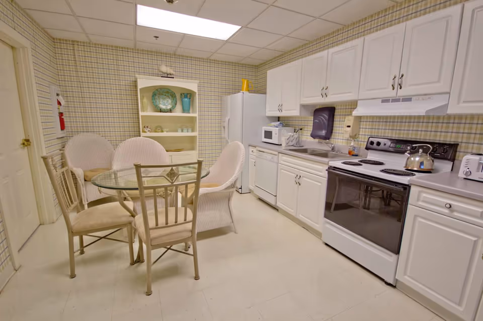 Small communal kitchen with white cabinets and appliances, a round glass dining table and cushioned chairs.