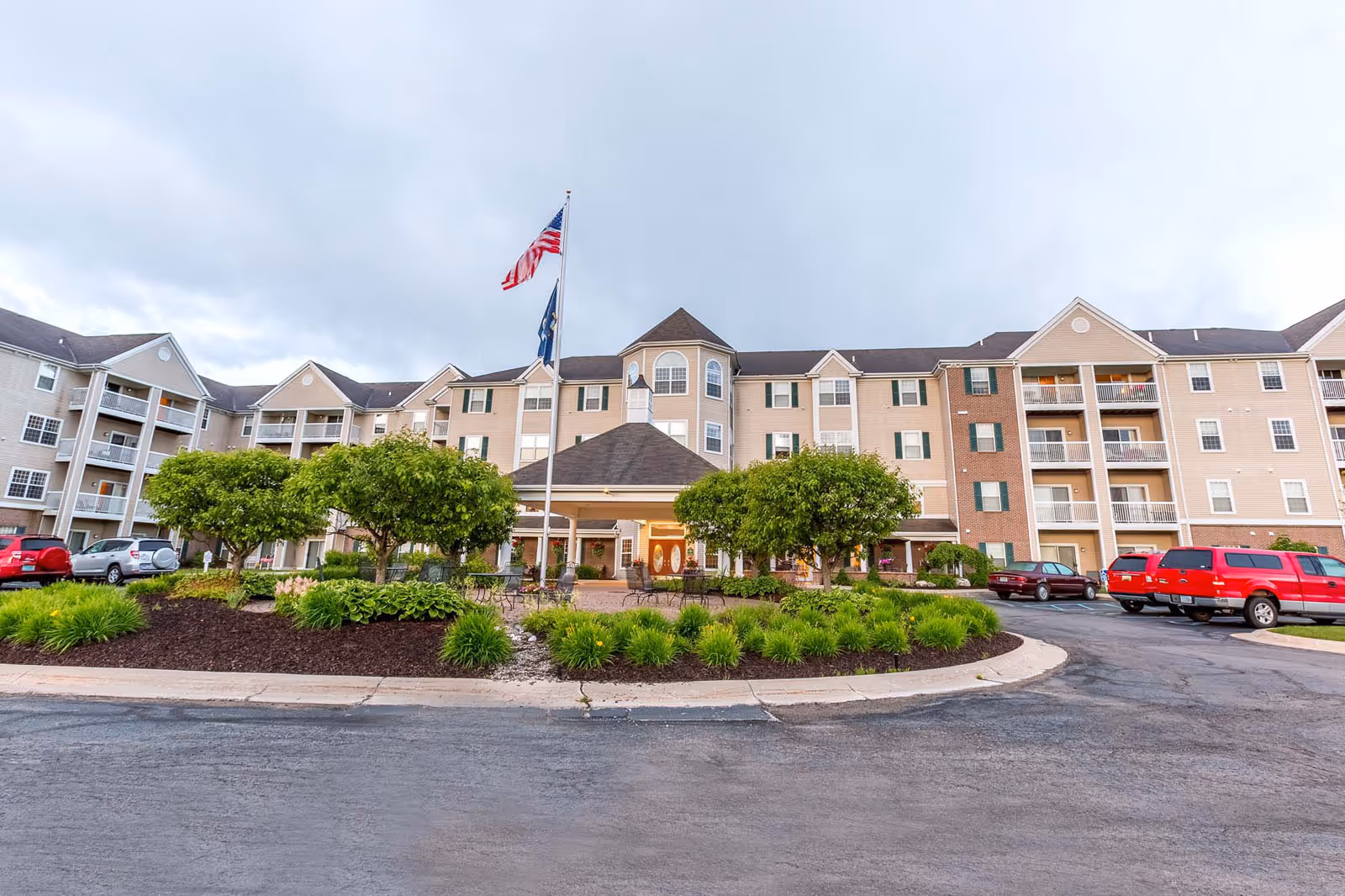 Front exterior of a multi-story senior living building with a covered entrance, flags, landscaped island, and parked cars.
