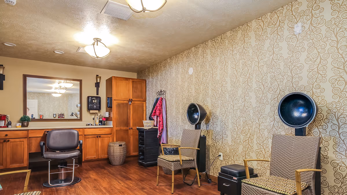 Interior salon room with a styling chair and mirror, wooden cabinets, patterned wallpaper, and two hooded hair dryers with seating.