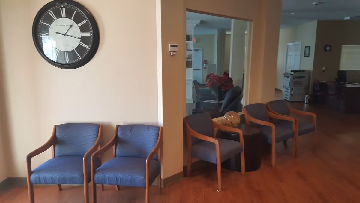 Interior view of a waiting area with six blue cushioned chairs with wooden armrests arranged along the walls. A large round wall clock with Roman numerals is mounted on a beige wall above two chairs. The floor is wooden, and there is a small round table with a decorative item between two chairs. In the background, there is an office area with desks, chairs, and office equipment.