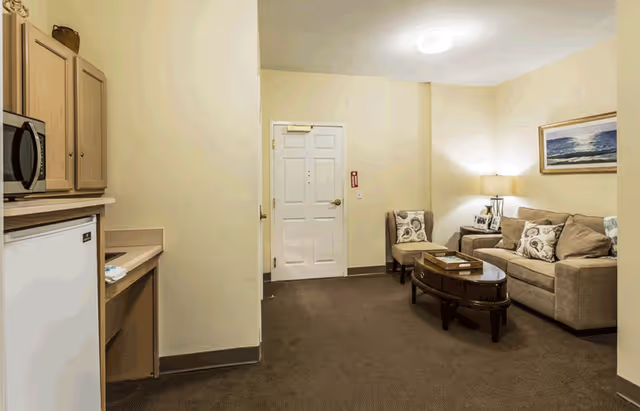 Interior view of a living area in an assisted living facility showing a small kitchenette with a microwave and mini fridge on the left, and a sitting area with a beige sofa, two chairs, a coffee table, a lamp, and a framed picture on the wall. A white door is visible in the background.