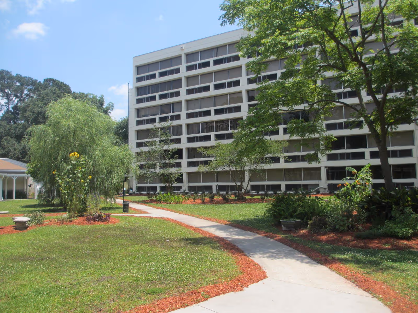 A multi-story residential building with numerous windows, surrounded by a well-maintained garden with green grass, trees, and flower beds. A concrete pathway curves through the garden leading towards the building under a partly cloudy blue sky.
