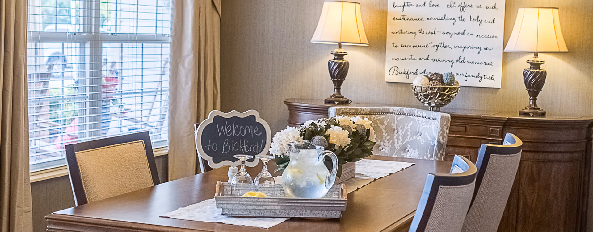 A warmly lit dining room with a wooden table set with a tray holding a pitcher of water with lemon slices, glasses, and a small chalkboard sign that says 'Welcome to Bickford.' The table is decorated with a floral centerpiece and a lace runner. Two table lamps with beige shades sit on a sideboard against the wall, which also has a framed inspirational quote hanging above it. Beige curtains frame a window with white blinds.