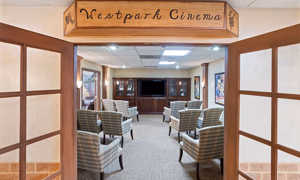 View through double wooden doors into a small cinema room named Westpark Cinema with eight plaid upholstered chairs arranged in two rows facing a large flat-screen TV mounted on a wooden cabinet. The room has beige walls, carpeted floor, and movie posters on the walls.