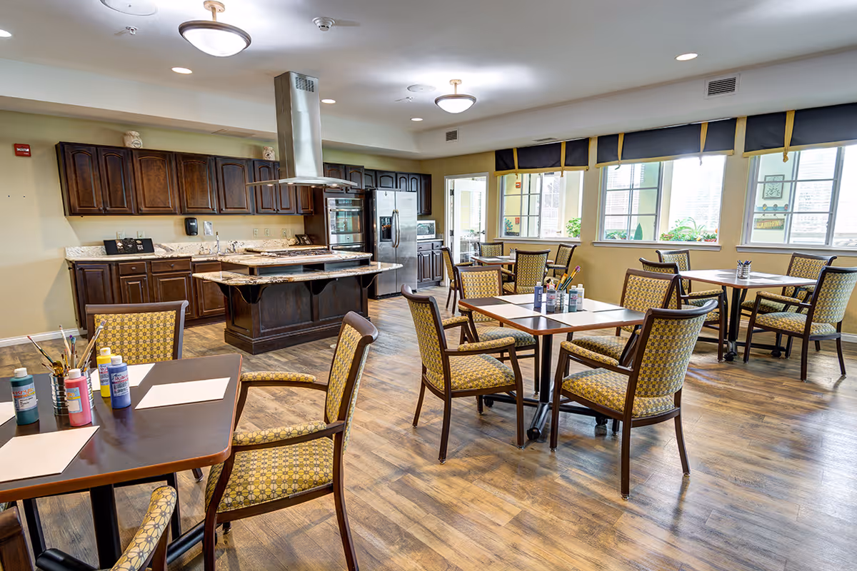 A bright and spacious room in a senior living facility featuring a large kitchen area with dark wooden cabinets, a central island with a range hood, and stainless steel appliances. Several tables with patterned cushioned chairs are arranged around the room, some with art supplies like paint bottles and brushes on them. Large windows let in natural light, creating a welcoming atmosphere.