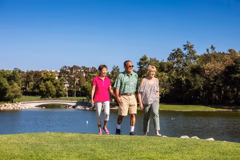 Three seniors walking together on a grassy area near a pond with trees and a small bridge in the background under a clear blue sky.