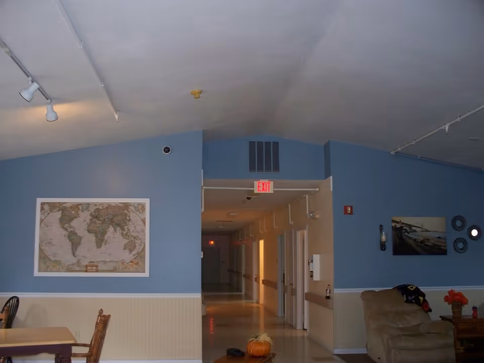 Interior view of a senior living facility hallway with blue and beige walls. On the left side, there is a dining area with a wooden table and chairs, and a world map hanging on the wall. On the right side, there is a beige armchair with a colorful blanket draped over it, a small table with a vase of flowers, and wall decorations including a photo and circular mirrors. The hallway extends into the distance with several doors and an illuminated exit sign above the doorway.