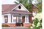 Front entrance of a brick senior living building with a covered portico supported by white columns and surrounding landscaping.