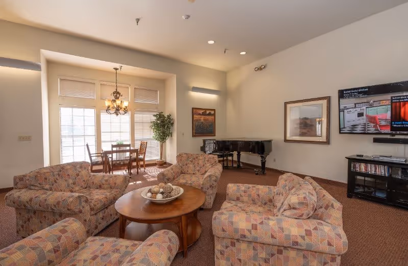 A senior living facility common area with patterned upholstered armchairs and sofas arranged around a wooden coffee table. In the background, there is a dining table with chairs near large windows with blinds, a chandelier hanging above, a grand piano, framed artwork on the walls, and a wall-mounted flat-screen TV with a media console below.