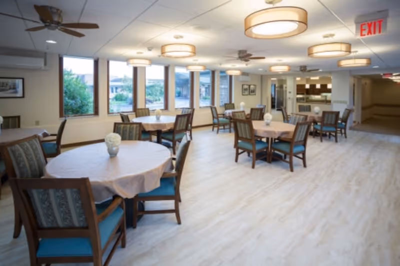 Spacious dining room with round tables draped in tablecloths, wooden chairs, pendant lights, and large windows looking out to greenery.