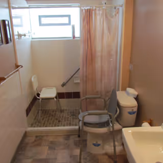 Bathroom with a walk-in shower featuring a white shower chair and a grab bar. There is a toilet with a gray shower chair placed over it, a sink, and a window above the shower area. The walls are tiled halfway up and painted beige above.