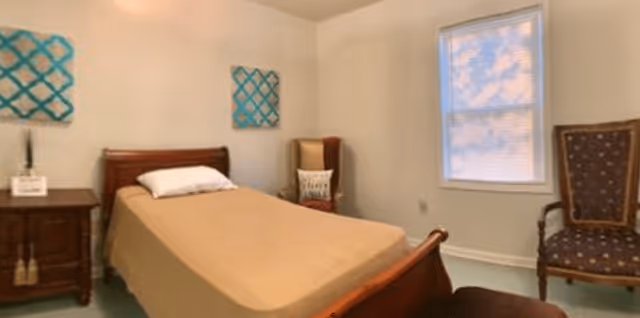 A simple bedroom with a single bed covered with a beige blanket and a white pillow. There is a wooden nightstand with a small plant and a framed item on it. Two decorative blue wall hangings are above the bed. The room has two upholstered chairs, one with a pillow, and a window with blinds partially closed.
