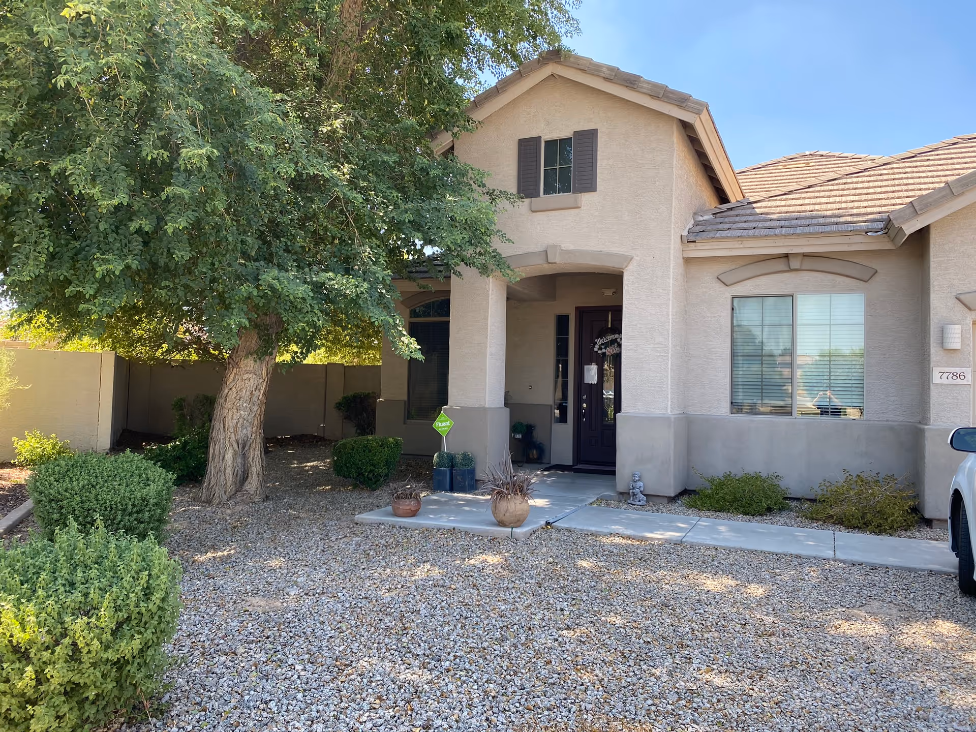 Front exterior view of a single-story house with beige stucco walls and a tiled roof. There is a large tree and several bushes in the gravel-covered front yard. A concrete pathway leads to a covered front porch with a dark-colored door and a welcome sign. A white vehicle is partially visible on the right side of the image.