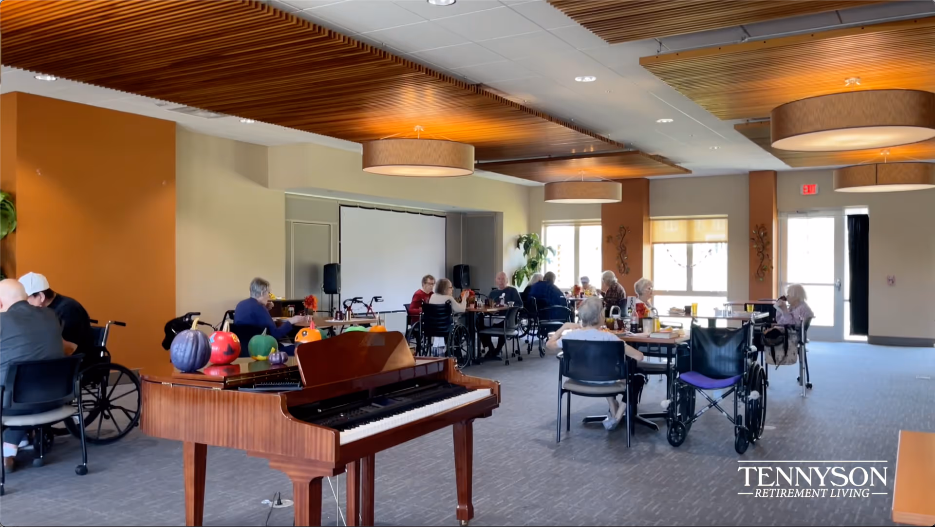 A spacious common area in an assisted living facility with elderly residents sitting at tables, some in wheelchairs, engaging in conversation and activities. The room features a wooden piano decorated with colorful pumpkins, modern ceiling lights, and large windows letting in natural light.