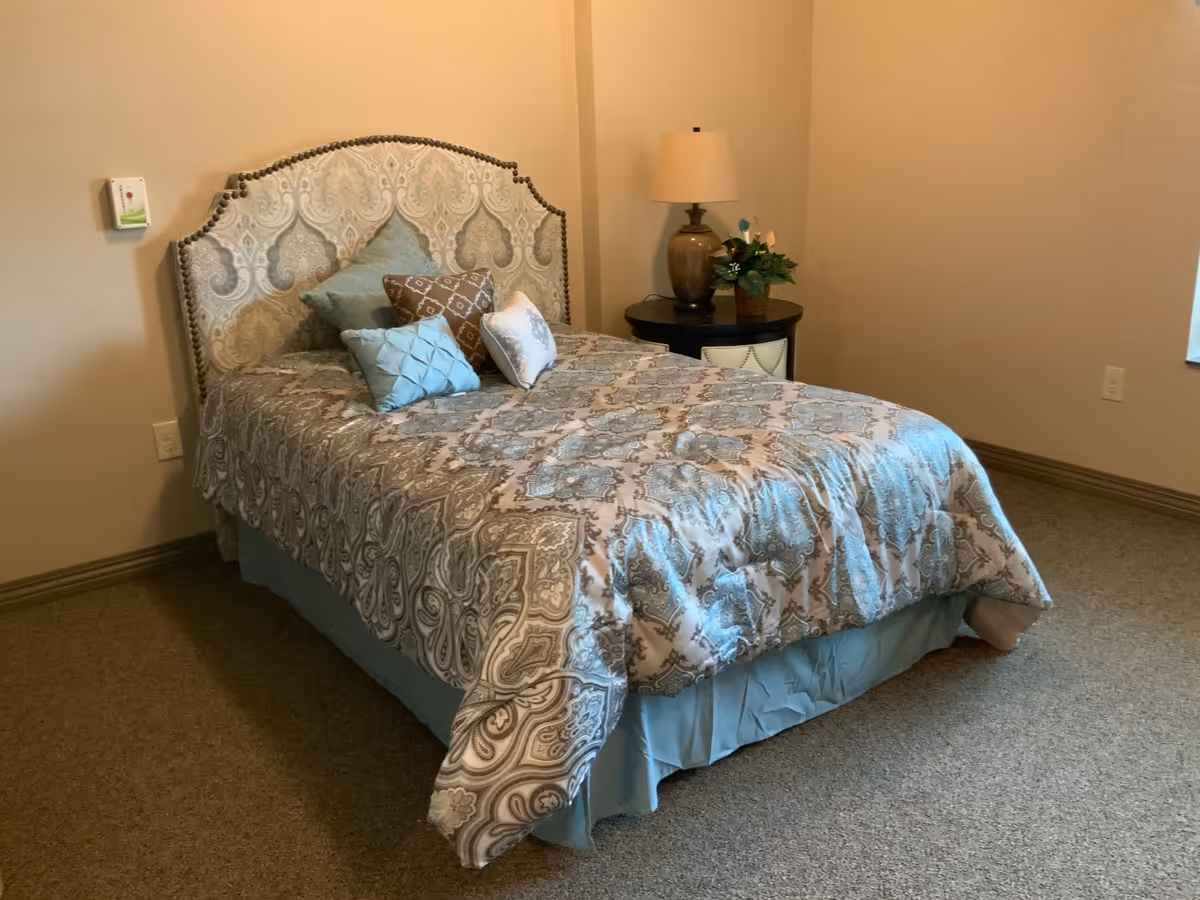 A neatly made bed with a patterned headboard and multiple decorative pillows in a simple bedroom. Next to the bed is a round nightstand with a lamp and a small plant. The room has beige walls and carpeted floor.