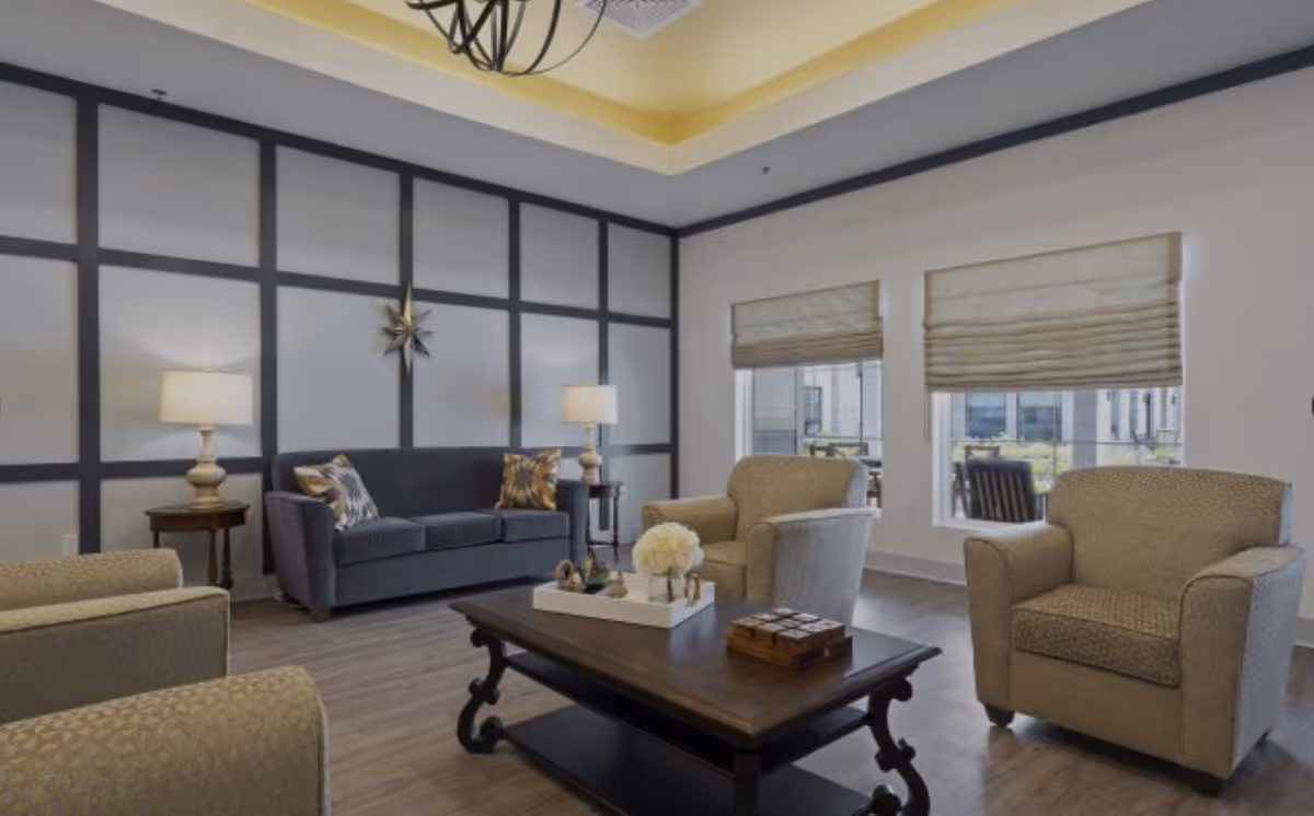 A cozy living room with a dark blue sofa, two beige armchairs, a wooden coffee table with decorative items, two side tables with lamps, large windows with beige Roman shades, and a wall decorated with black grid paneling and a star-shaped ornament.