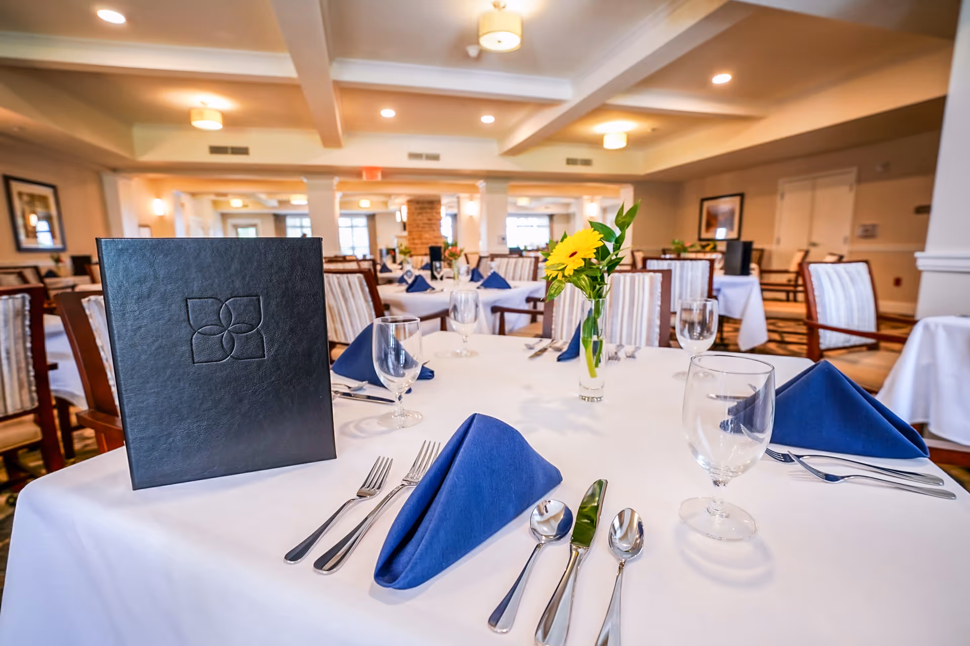 A neatly set dining table in a spacious dining room with white tablecloth, blue folded napkins, silverware, water glasses, a black menu, and a small vase with a yellow flower. The room has multiple tables and chairs, warm lighting, and framed artwork on the walls.