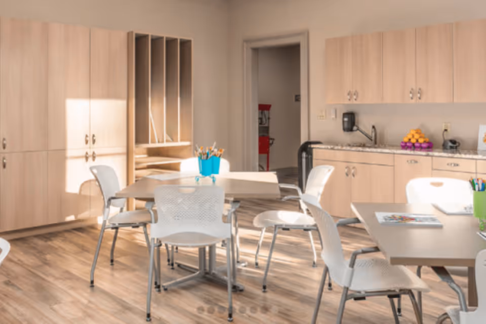 A bright room with light wood cabinets and countertops, several tables with white chairs around them, and containers holding colored pencils and art supplies, suggesting a creative or activity space.