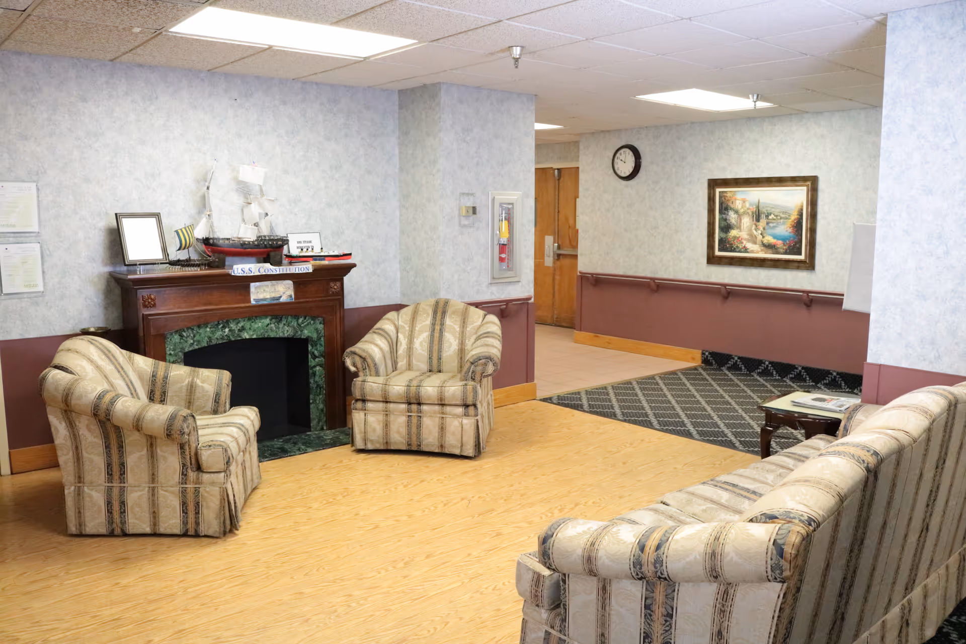 A cozy sitting area in a senior living facility with three upholstered armchairs and a matching sofa arranged around a decorative fireplace. The fireplace mantle holds a model ship and framed pictures. The room has light-colored walls with a maroon wainscoting, a wooden floor, a wall clock, and a framed landscape painting. There is a patterned rug near a doorway leading to another area.