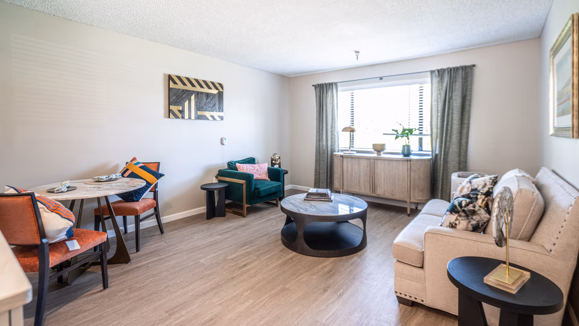 Sunlit living room with a sofa, armchair, round coffee table, small dining table and a window with curtains.