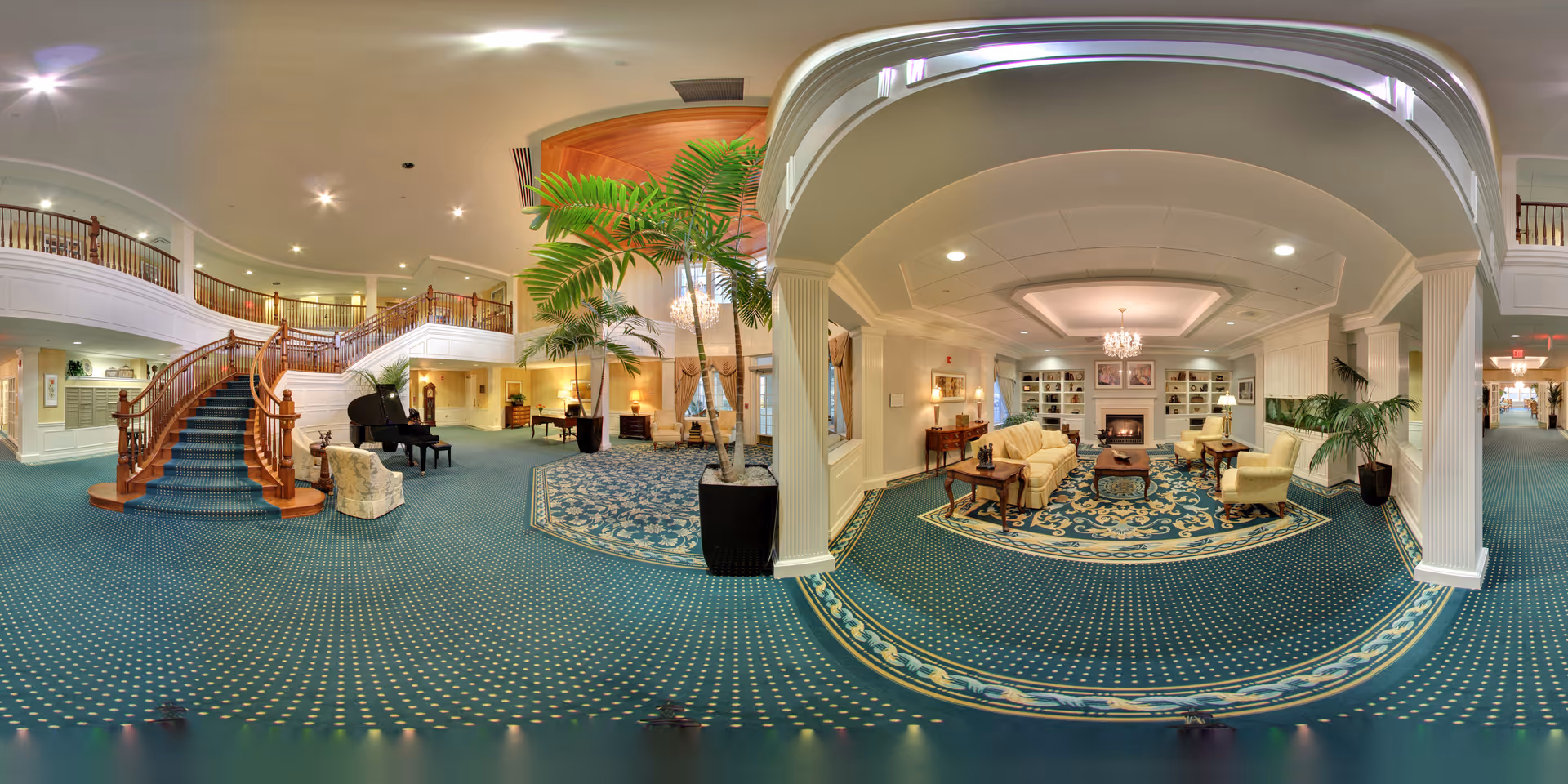 Spacious senior living lobby with a curved wooden staircase, piano, plants, and a seating area with a fireplace under chandeliers.