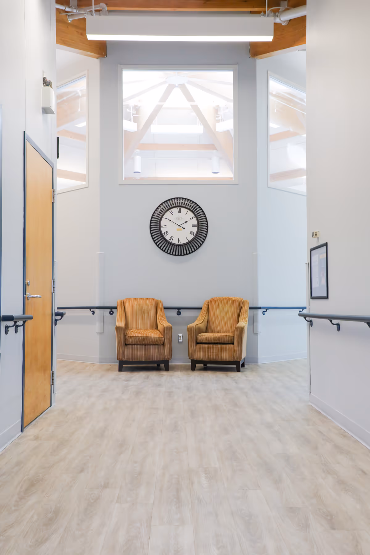 A bright hallway in a senior living facility with light wood flooring, white walls, and a high ceiling with exposed wooden beams. Two orange upholstered armchairs are placed side by side against the wall beneath a large round clock with Roman numerals. Handrails run along both sides of the hallway, and there is a wooden door on the left side.