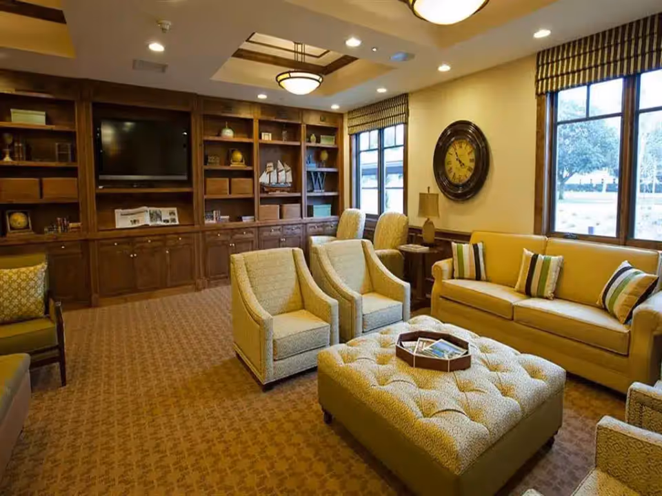 Comfortable senior living lounge with sofas, armchairs, a tufted ottoman, built-in wooden shelving and large windows.