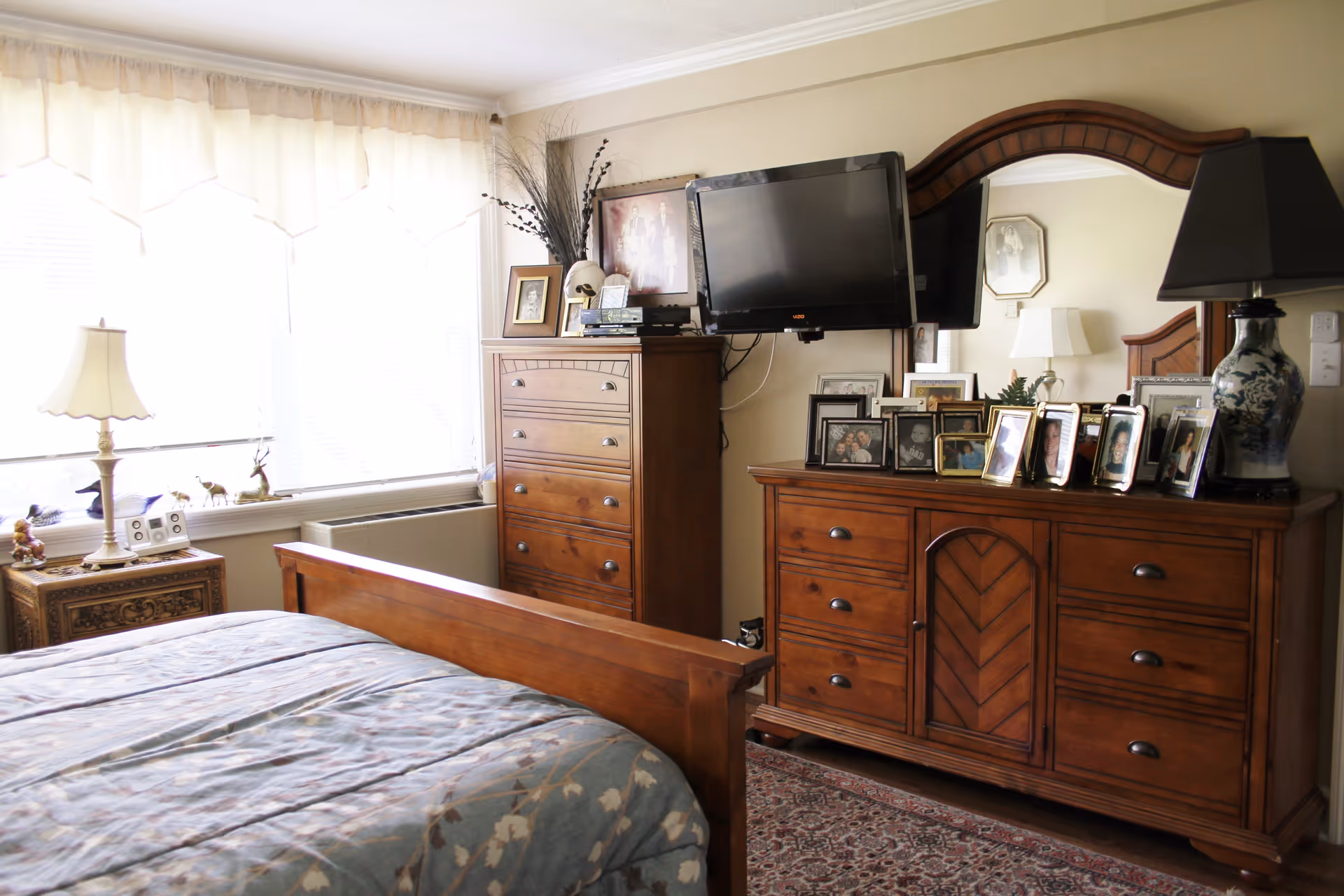 A bedroom with wooden furniture including a bed, a tall dresser, and a wide dresser with a mirror. The dresser is decorated with numerous framed photos and a large lamp. A flat-screen TV is mounted on the wall above the tall dresser. The room has a large window with sheer curtains allowing natural light to fill the space.