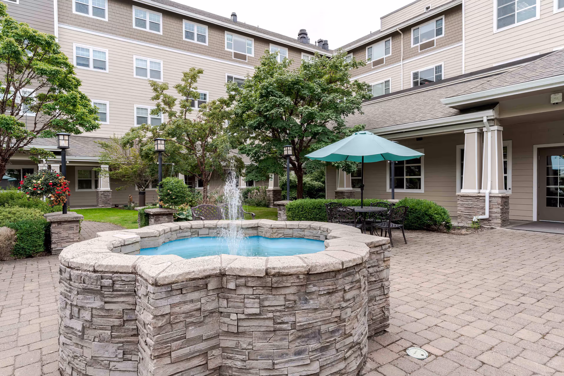 Outdoor courtyard area of a retirement community featuring a stone water fountain in the center, surrounded by paved walkways, green trees, bushes, and outdoor seating with a table and umbrella. The multi-story beige building with many windows forms the background.