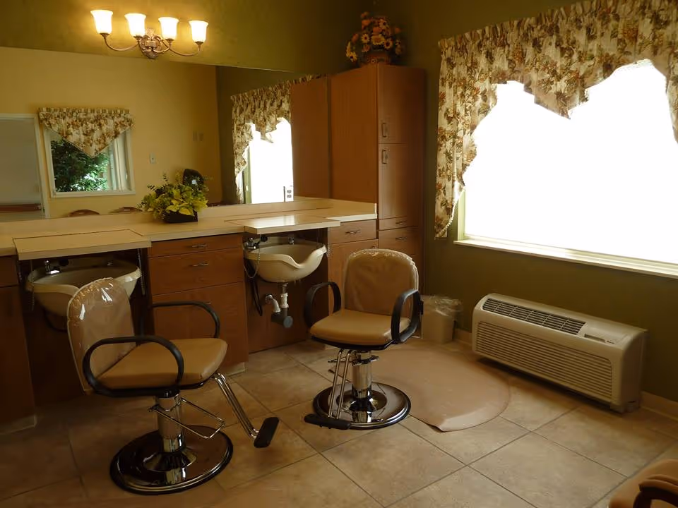 Interior of a salon area with two beige salon chairs in front of a counter with sinks. The room has floral curtains on the windows, a wall-mounted air conditioning unit, and a chandelier light fixture on the ceiling. There is a large mirror on the wall behind the counter and a floral arrangement on top of a cabinet.