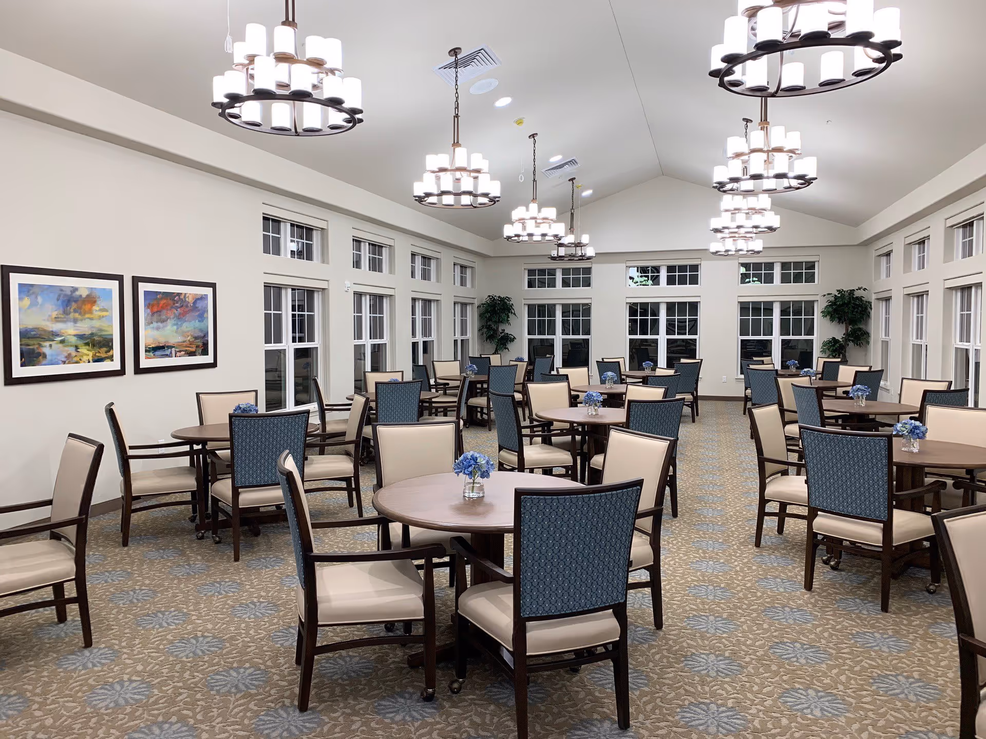 A spacious dining room with multiple round wooden tables, each surrounded by chairs with beige seats and blue patterned backs. The room has large windows along the walls, beige walls, and a patterned carpet with blue and beige tones. Several modern chandeliers with multiple lights hang from the ceiling, and there are two framed colorful paintings on one wall and potted plants in the corners.