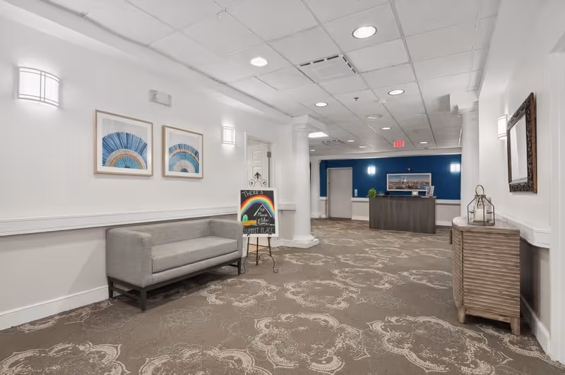 Interior hallway of a senior living facility with patterned carpet, white walls, and ceiling lights. On the left side, there is a gray cushioned bench beneath two framed blue and beige artworks. A small chalkboard sign with a rainbow drawing and the words 'There's peace, relax, believe, Summit Place' is placed near the bench. At the end of the hallway, there is a reception desk against a blue accent wall with a framed picture above it. On the right side, there is a wooden cabinet with decorative lanterns and a framed mirror above it.
