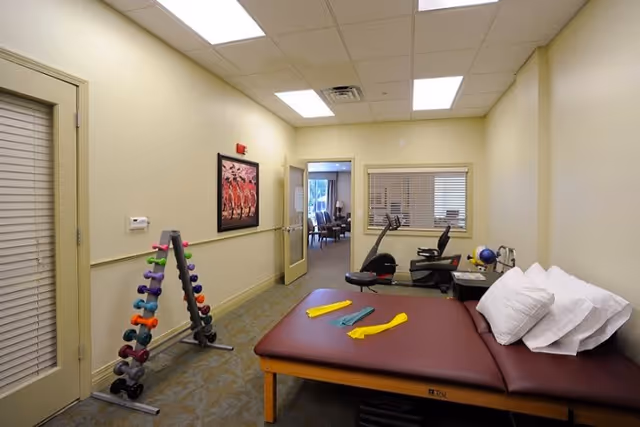 A therapy or exercise room with a padded treatment table holding resistance bands and pillows, a rack of colorful dumbbells, and two exercise bikes near a window. The room has beige walls, a carpeted floor, and a door leading to another area with chairs visible.