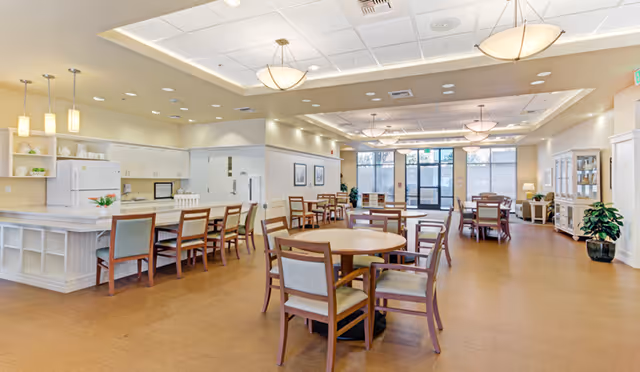 A spacious and well-lit dining area with multiple round and rectangular wooden tables surrounded by chairs. The room features a kitchen area with white cabinets, a refrigerator, and a microwave on the left side. Large windows and glass doors at the far end allow natural light to fill the space. The ceiling has recessed lighting and hanging light fixtures, and there are plants and decorative items placed around the room.