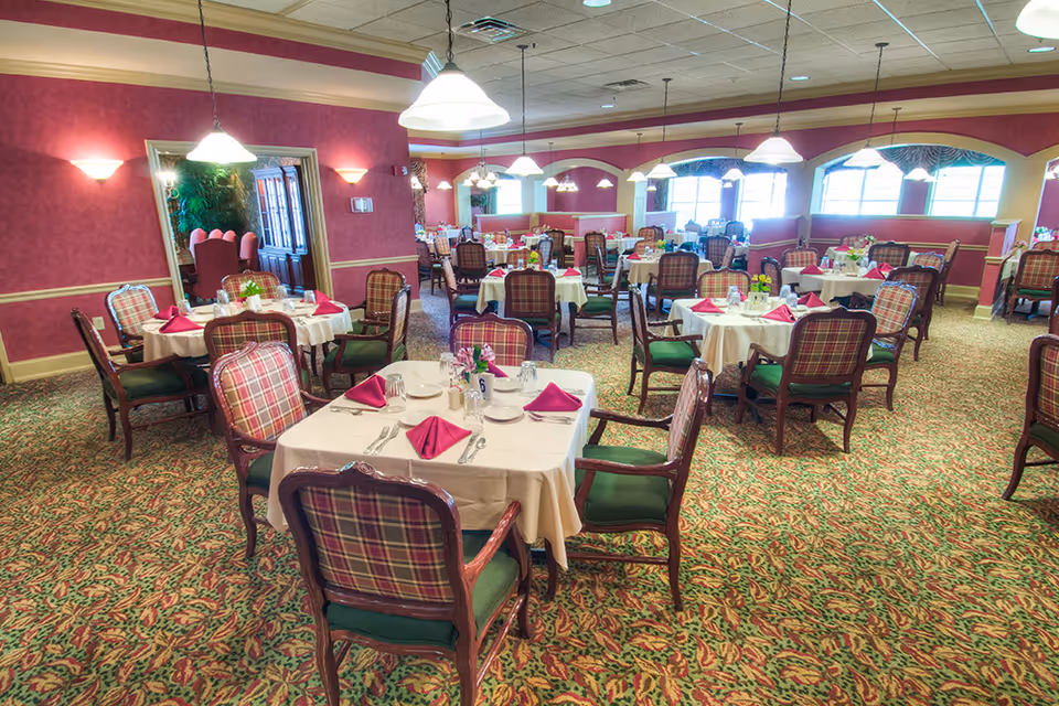 A spacious dining room with multiple round tables covered with white tablecloths, each set with plates, glasses, silverware, and folded red napkins. The chairs have wooden frames with plaid upholstery and green cushions. The room has patterned carpet, red walls with beige trim, and several hanging pendant lights. Large windows allow natural light to enter the room.