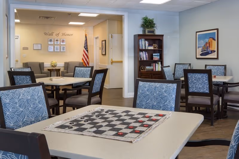 A common area in a senior living facility with several tables and chairs, one table in the foreground has a checkers board with red and black pieces. In the background, there is a 'Wall of Honor' with framed photos and an American flag. A bookshelf with books and decorative items is against the wall, and a framed picture hangs on the right wall.