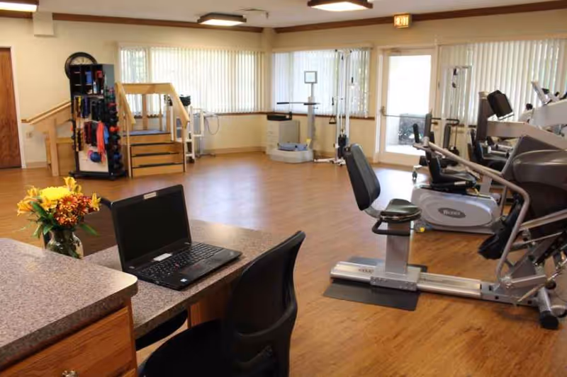 Bright fitness/therapy room with exercise machines, a desk with a laptop and flowers, and shelving with weights.