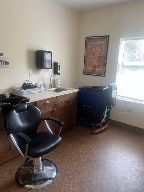 A small room with a black salon chair in front of a countertop with a sink. On the countertop are some hair care items and a hair washing basin. A large window lets in natural light, and a framed poster hangs on the wall. A cleaning machine is positioned near the window.