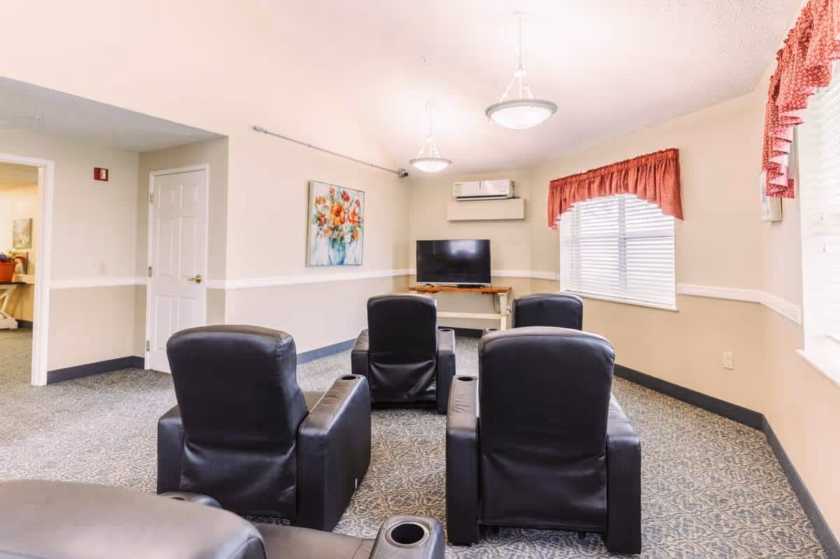 A small common area in a senior living facility with four black leather recliners arranged in two rows facing a flat-screen TV mounted on a wooden stand. The room has beige walls with white trim, a patterned carpet, two hanging ceiling lights, a window with red valance curtains, and a floral painting on the wall.